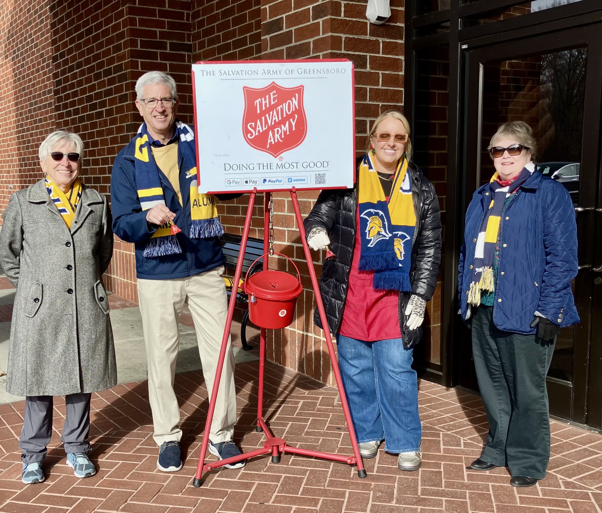 Four alumni ringing a bell around a Salvation Army donation area.